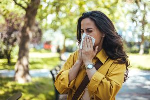 Portrait of unhealthy cute female in yellow top with napkin blowing nose